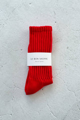 A single bright red ribbed ballet sock with a white paper band label reading 'LE BON SHOPPE' and 'BALLETS SOCKS', laid flat on a light textured gray surface.