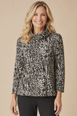A smiling blonde woman wearing a black and white abstract patterned long-sleeve blouse with a mock neck, paired with black pants, against a solid beige background.
