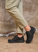 Person wearing black shoes and beige pants against a textured wall.