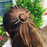 Person with a gold hair accessory in their brown hair, outdoors with greenery in the background