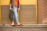 Person wearing brown shoes standing on a sidewalk with a yellow shutter and brick wall in the background
