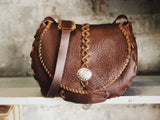 Brown leather bag with braided strap and decorative button on a wooden surface.