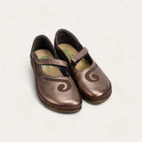 A pair of metallic bronze Mary Jane shoes with a decorative swirl design on the toe, displayed on a light off-white background.