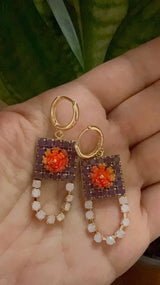 Gold hoop earrings with purple and orange floral designs held in a hand against a green leafy background.