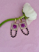A pair of gold drop earrings with square crystal-encrusted frames, purple flower centers, and dangling rows of dark blue and white stones, displayed on a pink background next to a white flower.