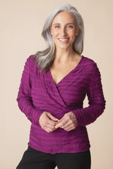 A smiling woman with gray hair wearing a textured, ruched long-sleeve wrap top in a vibrant magenta-purple color, paired with black pants, against a solid beige background.